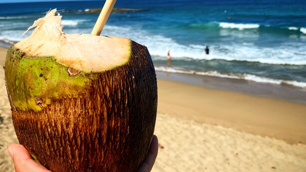 Una mano sosteniendo un coco con una pajita, sobre un fondo de playa y océano.