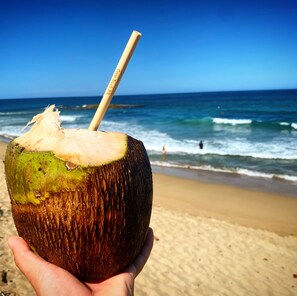 A hand holding a coconut with a straw, set against a beach and ocean backdrop. - Beachfront Apt w/ Pool & Private Access – Walk to Jobos & Shacks (Isabela)