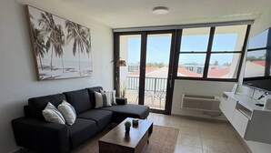 Una sala de estar moderna con un sofá negro, un sillón gris y una mesa de centro de madera. Se ve una ventana grande con vista a palmeras y un edificio.