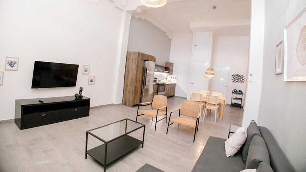 Una sala moderna con un televisor de pantalla plana, una mesa de centro de vidrio y un sofá. Se observan sillas de mimbre y una mesa de comedor al fondo.