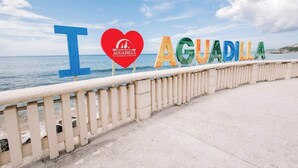 Vista costera con un cartel que dice "I ♥ AGUADILLA" y un logo en forma de corazón con el emblema de Coca-Cola.