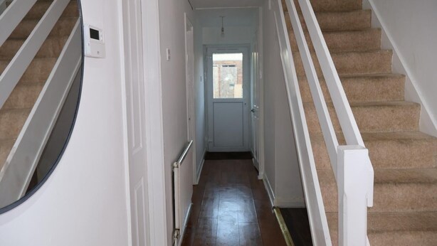 Un pasillo con una escalera alfombrada, paredes blancas y una puerta que da a una habitación luminosa.