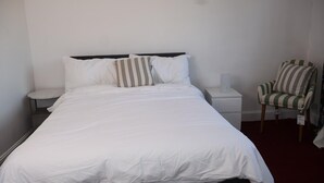 Una cama bien hecha con ropa de cama blanca y almohadones a rayas, una mesita de noche con una lámpara y un sillón a rayas.