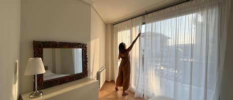 A hotel room with a large bed, a dresser, a mirror, and a window with sheer curtains.