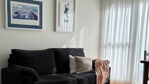Een woonkamer met een zwarte bank, twee ingelijste foto's aan de muur, en een groot raam met dunne gordijnen.