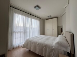Ein Schlafzimmer mit einem Bett, einem Kleiderschrank, einem Spiegel und einem Fenster mit durchsichtigen Vorhängen.
