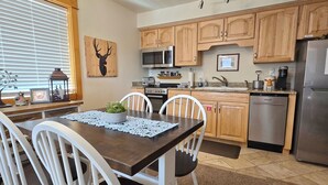 A modern kitchen with wooden cabinets, a stainless steel refrigerator, and a dining area with white chairs and a dark wooden table. - Elk Meadow Condo #6047 (Estes Park)