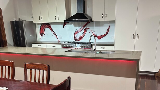 A modern kitchen with white cabinets, a central island with a red under-cabinet lighting, a stainless steel range hood, and a dining area with wooden chairs and a table. - Charming 3-bedroom cottage with AC in Torbanlea (Torbanlea)