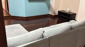 A living room with a sectional sofa, a flat-screen TV mounted on the wall, a small table with a tissue box, and a wooden floor. - Charming 3-bedroom cottage with AC in Torbanlea (Torbanlea)