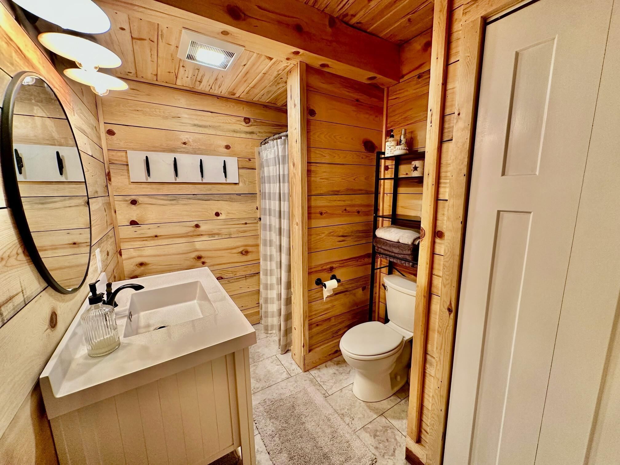 Une salle de bain aux murs lambrissés, avec un lavabo blanc, un miroir, des toilettes et une étagère contenant des articles de toilette.