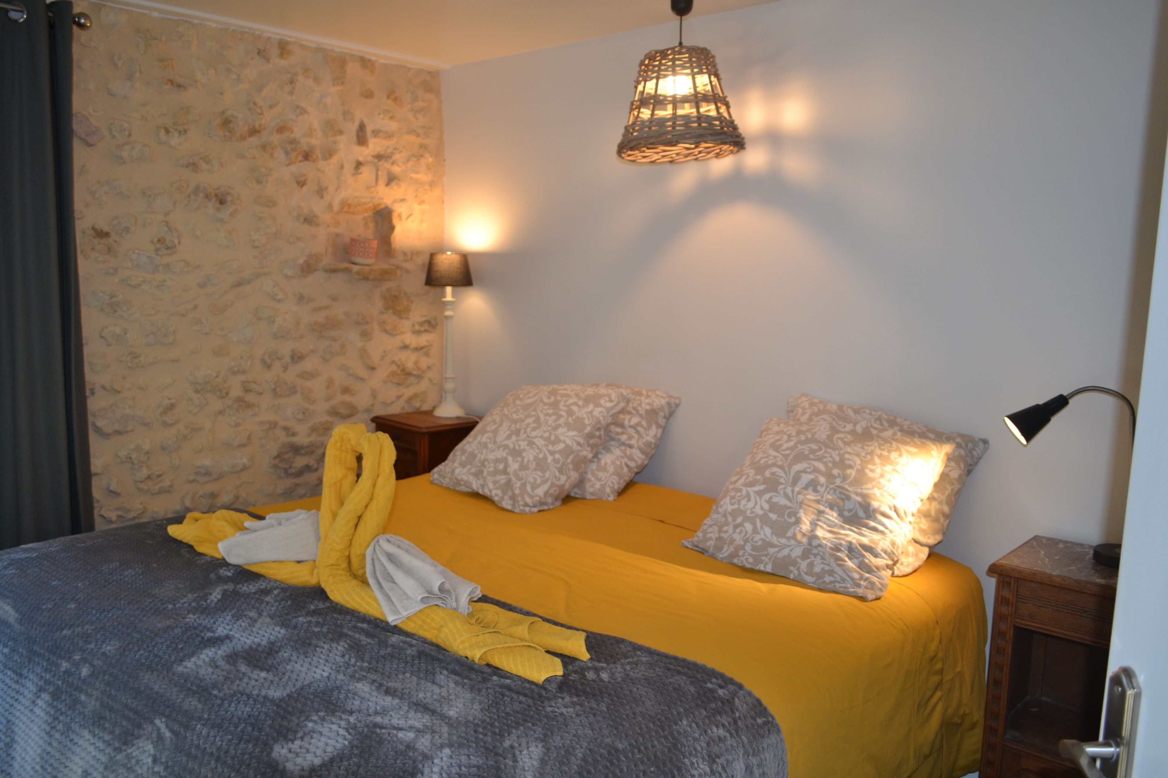 A bedroom with a stone wall, a bed with yellow bedding, a lamp, and a hanging light fixture.