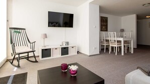 Ein Wohnzimmer mit Fernseher, einem Schaukelstuhl, einem Essbereich mit weißen Stühlen und einem kleinen Tisch mit Kerzen.