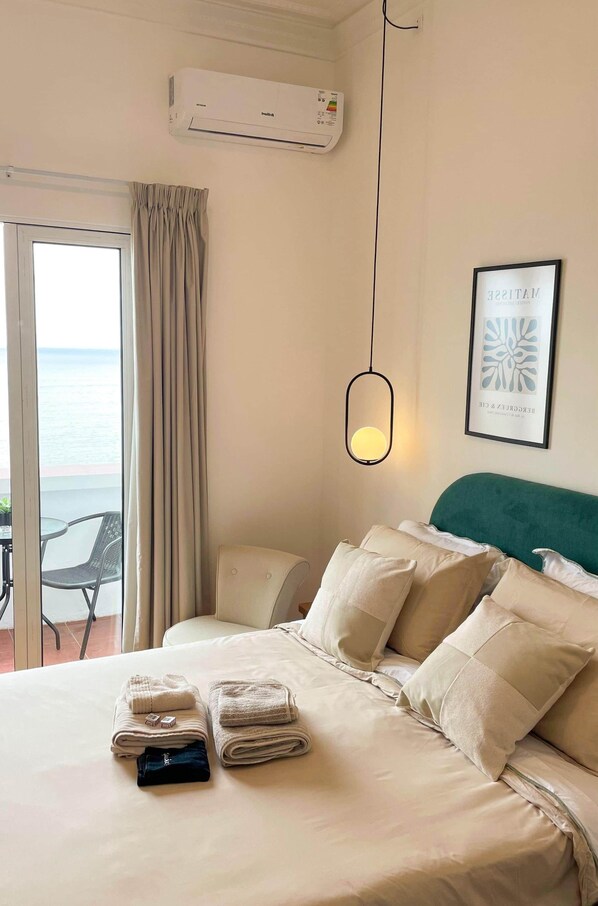 A hotel room with a bed, a view of the sea through a window, an air conditioning unit, and a framed artwork on the wall.