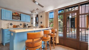 A kitchen with blue cabinets, a central island with stools, a coffee machine, and a large window with curtains. - Medano Beach, Marina, Downtown, Walkable, Cozy (Cabo San Lucas)