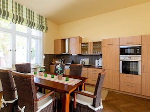 Een eetgedeelte met een houten tafel en rieten stoelen, een keuken met houten kastjes en apparatuur, en een raam met uitzicht op groen.