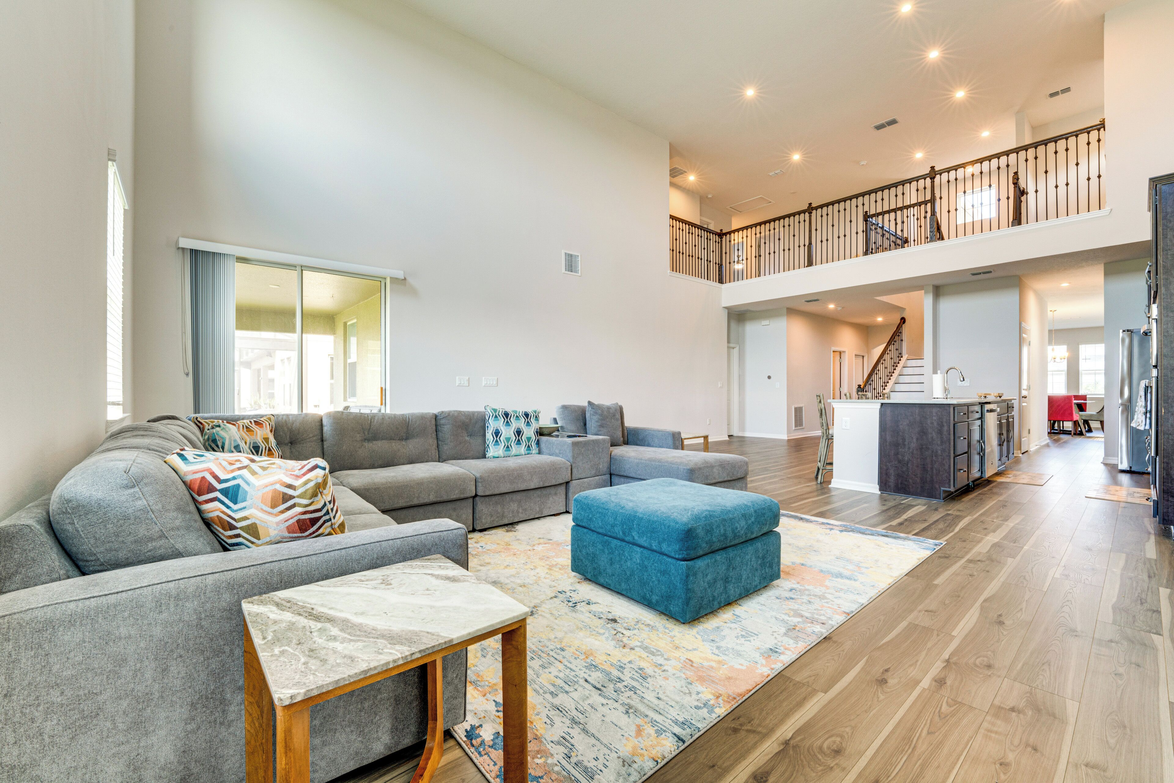 A spacious living area with a gray sectional sofa, a blue ottoman, and a wooden coffee table. A colorful rug is on the wooden floor.