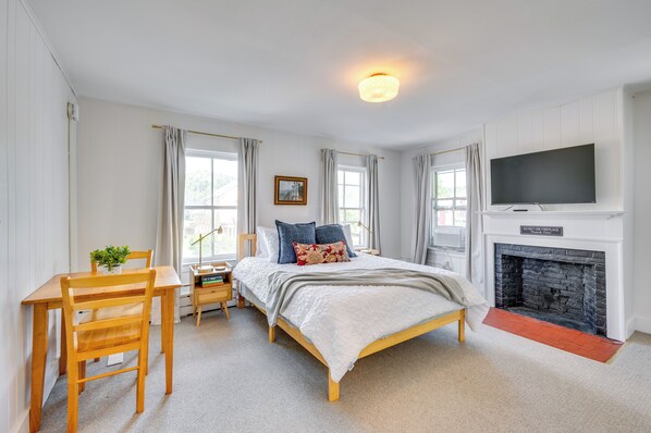 Une chambre à coucher avec un grand lit, un téléviseur fixé au-dessus d’une cheminée, un bureau avec une chaise et une fenêtre avec des rideaux.