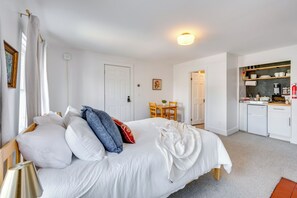 Une chambre comprenant un lit, une table de chevet, une lampe, une fenêtre avec des rideaux, une porte et un coin cuisine équipé d’un réfrigérateur, d’une cuisinière et d’étagères.