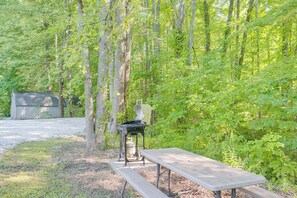 Une table de pique-nique et un barbecue sont installés dans un espace boisé, avec un abri de jardin en arrière-plan.