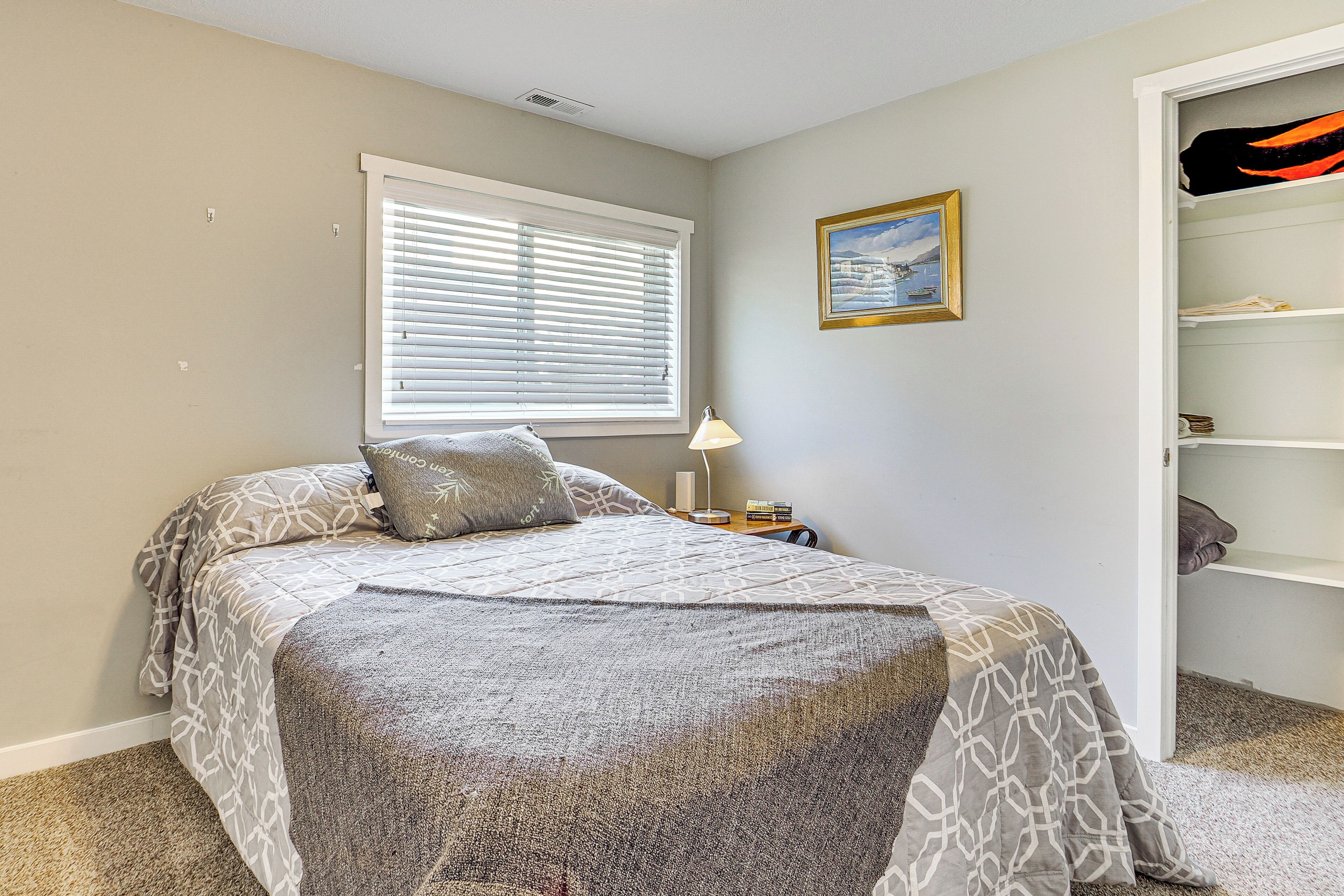 A bedroom with a bed, a nightstand, a lamp, a window with blinds, a framed picture on the wall, and a closet with shelves.