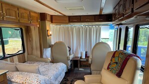 Un intérieur de camping-car accueillant, avec un lit, deux fauteuils, une petite table et une fenêtre avec des rideaux.