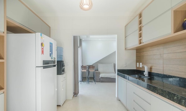 Eine moderne Küche mit weißen Schränken, einer schwarzen Arbeitsplatte und einer integrierten Spüle. Ein Wasserspender und ein Kühlschrank mit Gefrierfach sind vorhanden. Eine Treppe führt zu einem Schlafzimmer mit einem Bett und einem Sessel.