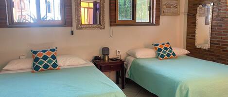 A room with two beds, each with a patterned pillow, a bedside table with a lamp, and a wall-mounted air conditioner.