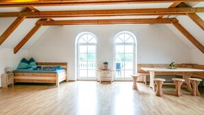 A spacious attic room with a wooden ceiling, two large windows, a wooden bench, a small table, and a wooden floor.