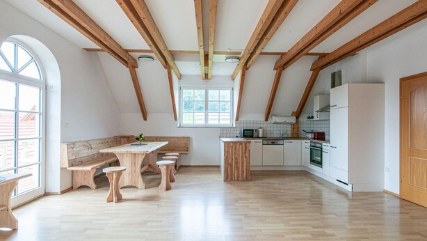 A spacious kitchen with wooden beams, a dining area with benches, and a large window providing natural light.