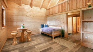 Ein Holzblockhaus im Inneren, mit einem Bett, einem kleinen Tisch mit Hocker und einer Tür zu einem anderen Raum.