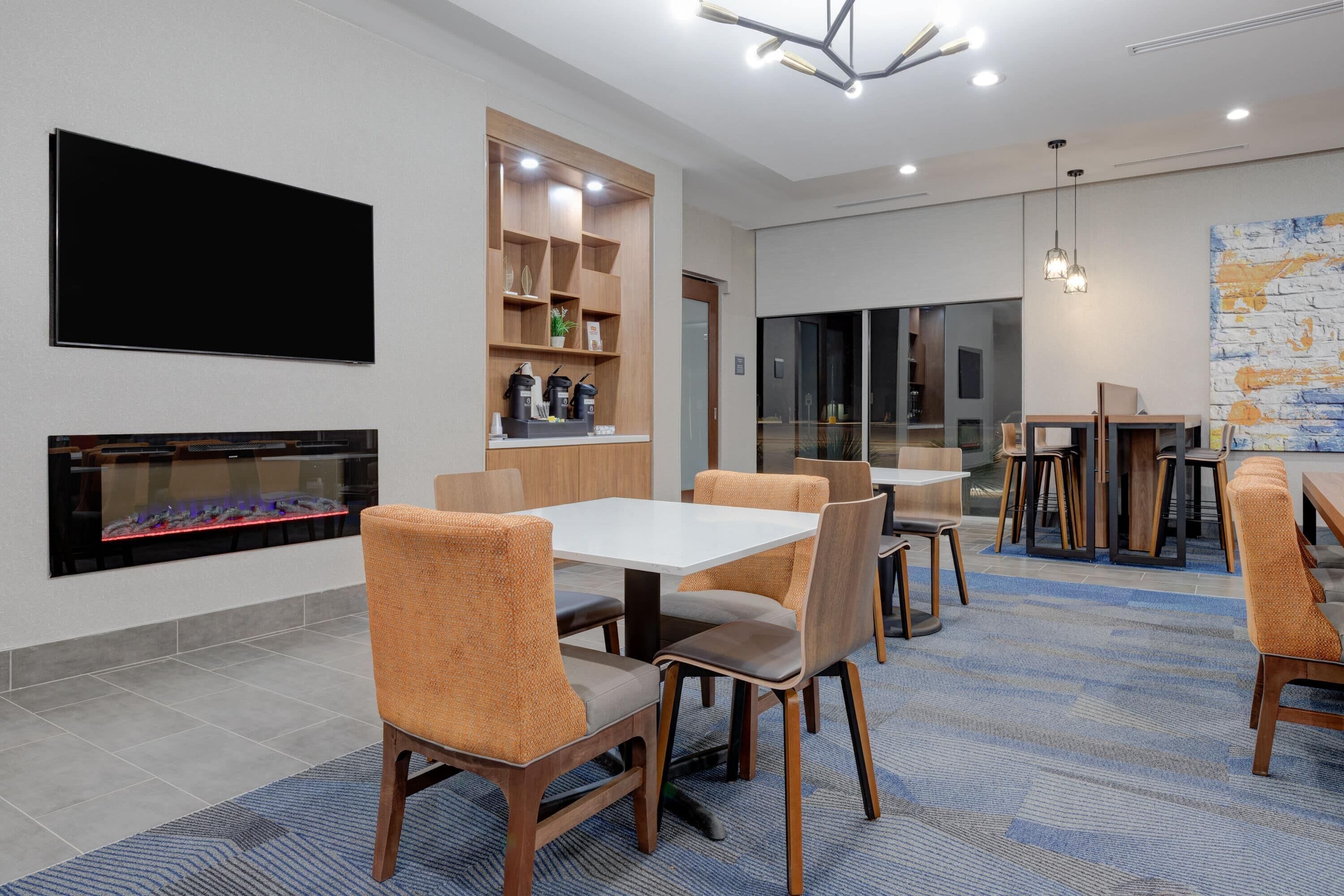 A modern hotel room with a large flat-screen TV, a fireplace, and a dining area with wooden furniture and a blue patterned carpet.