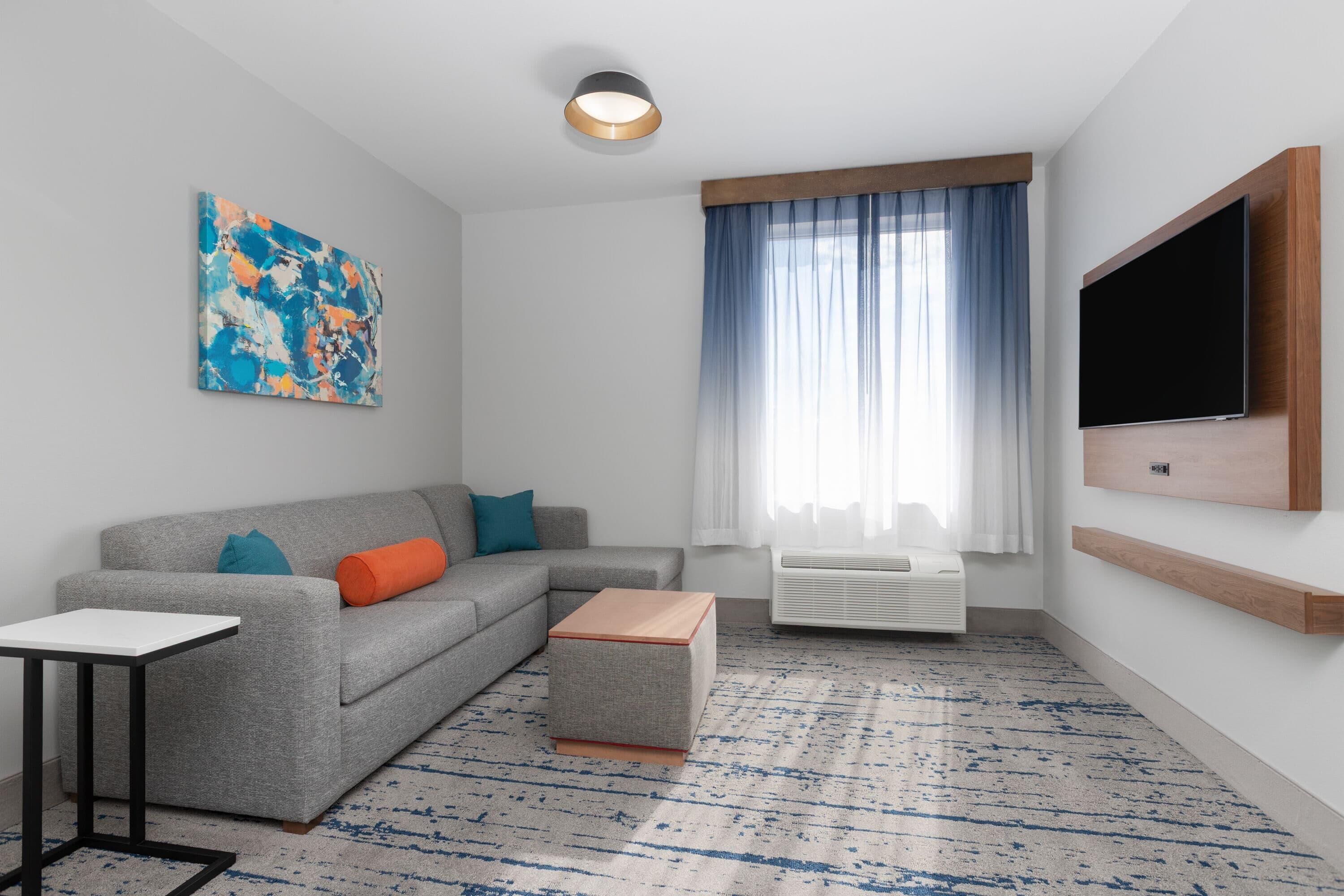 A modern living room with a grey sofa, a small wooden coffee table, a white side table, a large abstract painting, a flat-screen TV mounted on the wall, and a large window with blue curtains.