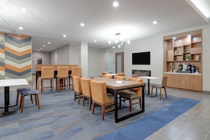 Um hall de hotel moderno com uma longa mesa de jantar, várias cadeiras, área de bar e uma televisão fixada na parede.