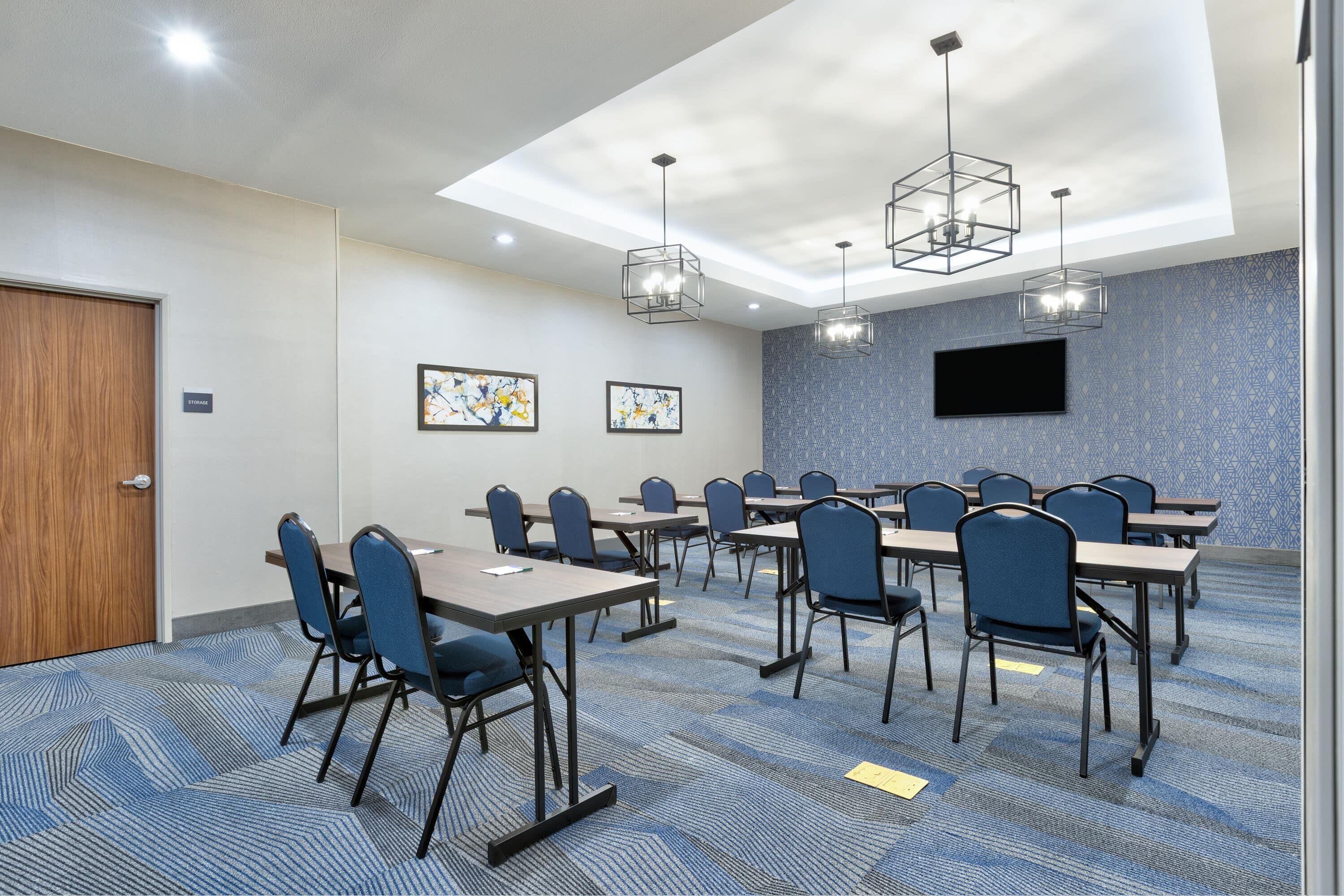 A conference room with a large screen, multiple tables, chairs, and geometric ceiling lights.