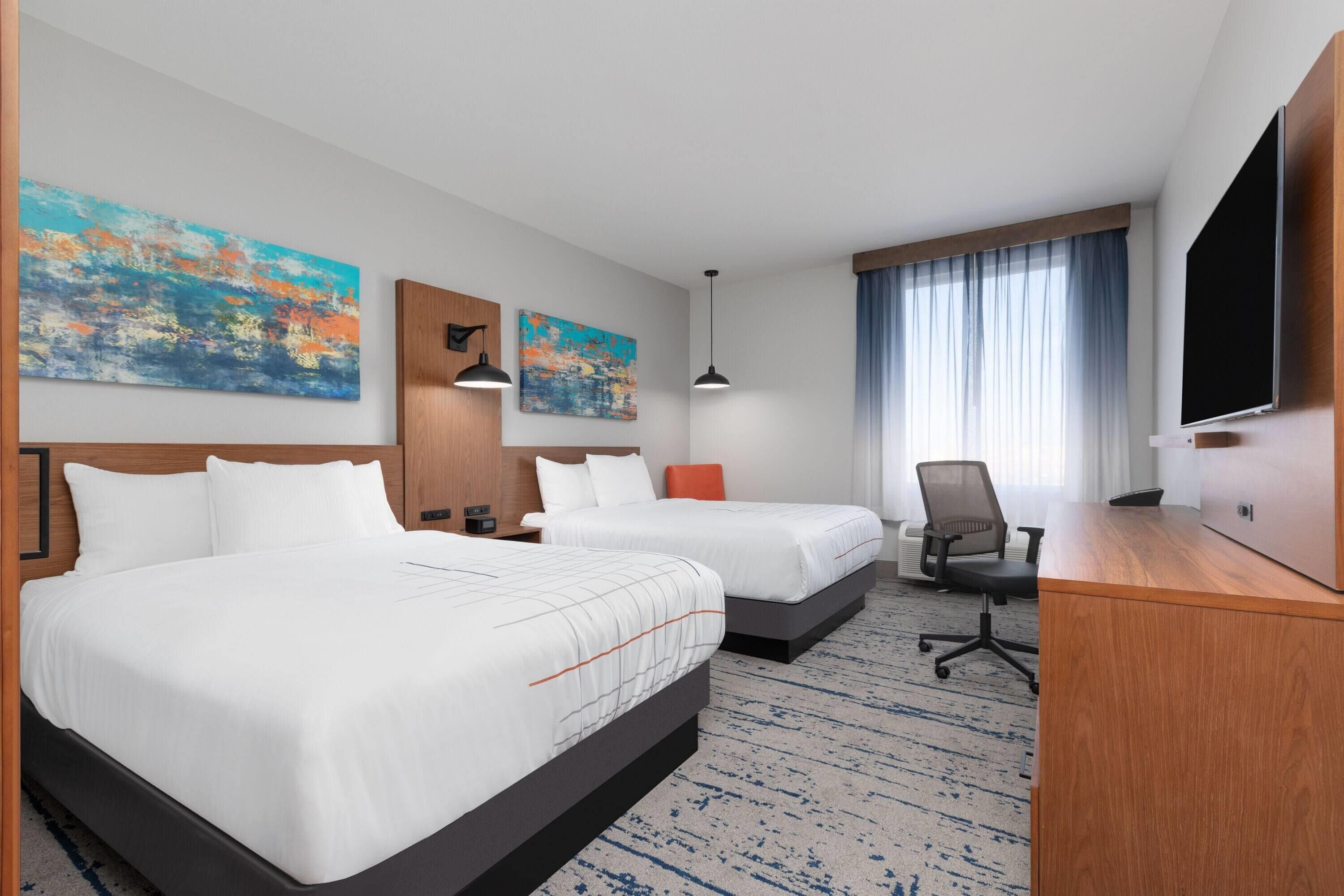 A hotel room with two beds, a desk, a TV, and artwork on the walls.