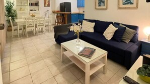 A living room with a dark blue sofa, a white coffee table with a book, and a dining area with a white table and chairs.