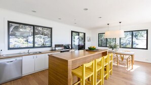 A modern kitchen with a central island, wooden cabinets, and large windows offering views of greenery. - Great Views, Walk To Beach and Bay In Bush Setting (KILLCARE)