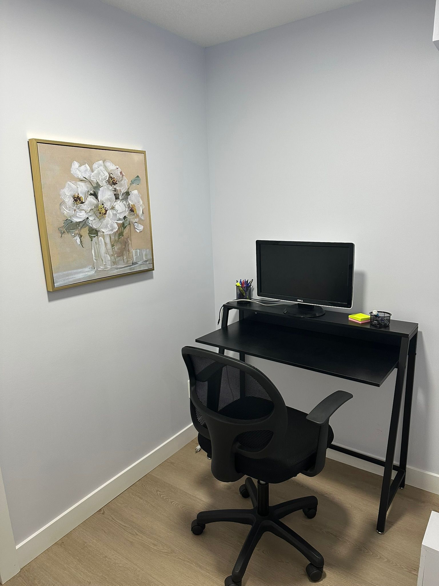 A room with a black desk, a black office chair, a Dell monitor, and a framed painting of flowers on the wall.
