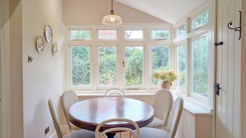 A dining area with a round table, four wicker chairs, a chandelier, and large windows with a view of greenery.