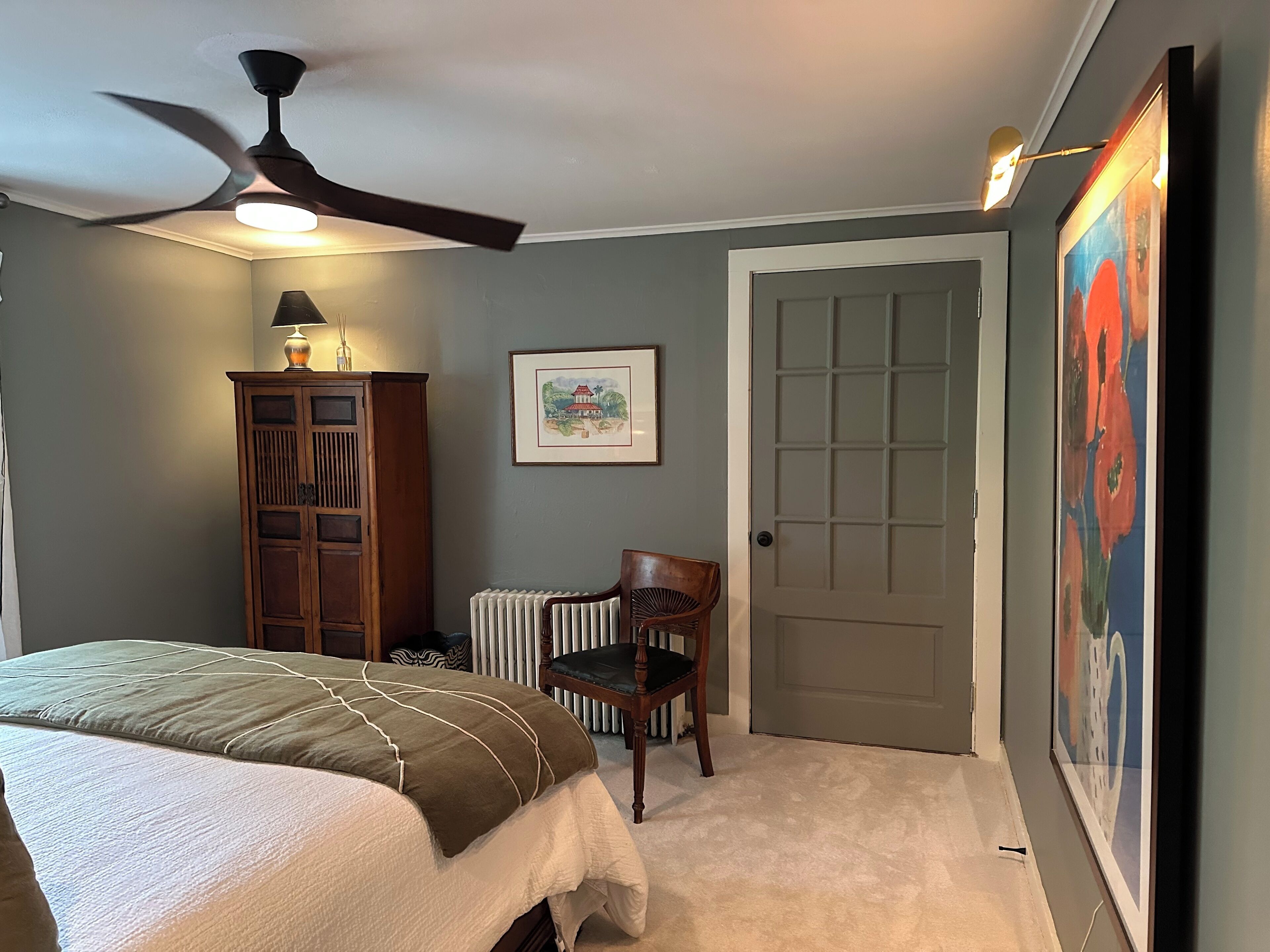 Une chambre à coucher avec un lit, un radiateur, une chaise, une porte et un ventilateur de plafond.