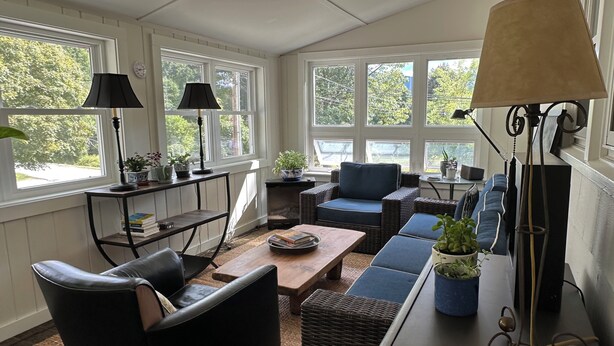 A cozy living room with a sofa, armchair, and coffee table, large windows with natural light, and potted plants.