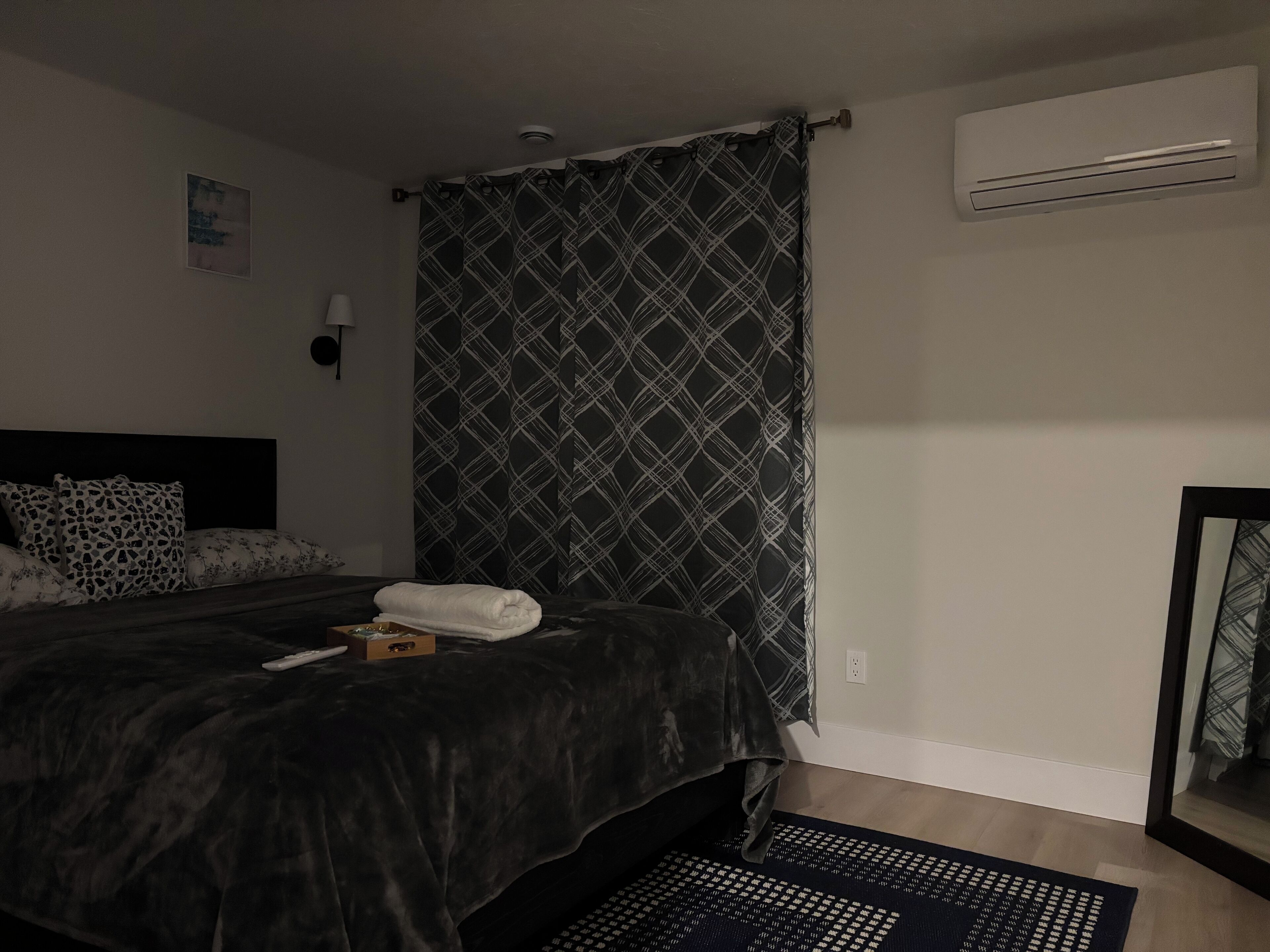Un dormitorio con cama, una colcha oscura, cortina estampada, aire acondicionado de pared y una alfombra pequeña.