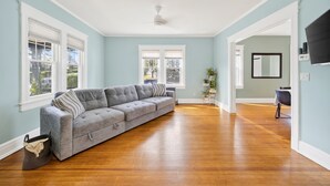 A spacious living room with a grey sectional sofa, wooden flooring, and large windows. - 16 Guest, 10 Bed, Family Getaway- 30 min from NYC (New Rochelle)