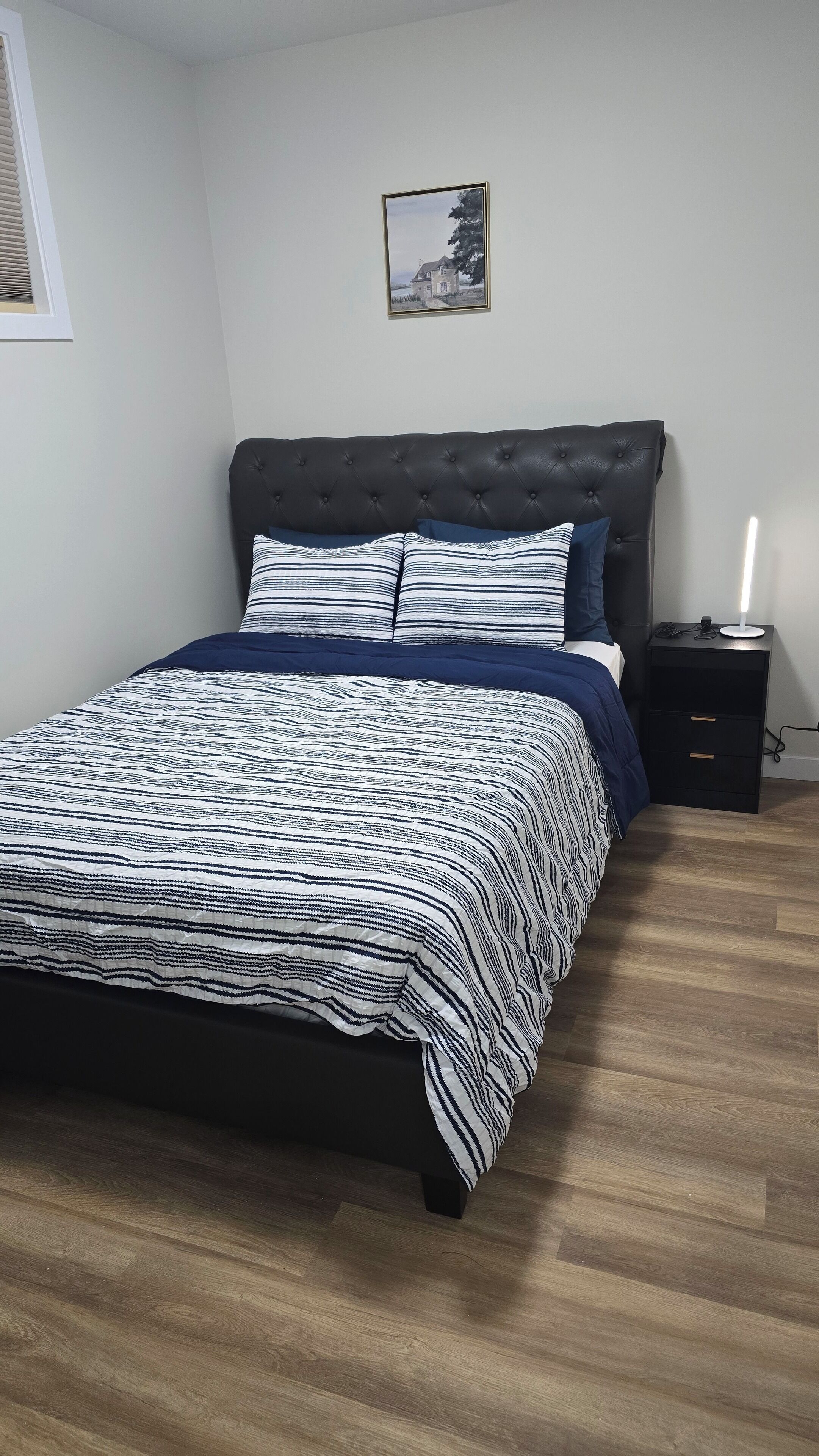 A bedroom with a dark headboard, a striped bedspread, a bedside table with a lamp, and a framed picture on the wall.