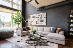 A modern living room with a sectional sofa, a glass coffee table, a large abstract painting, and a plant. - Obsidian | City Views, RL Therapy, Sauna & Lounge (Charlotte)