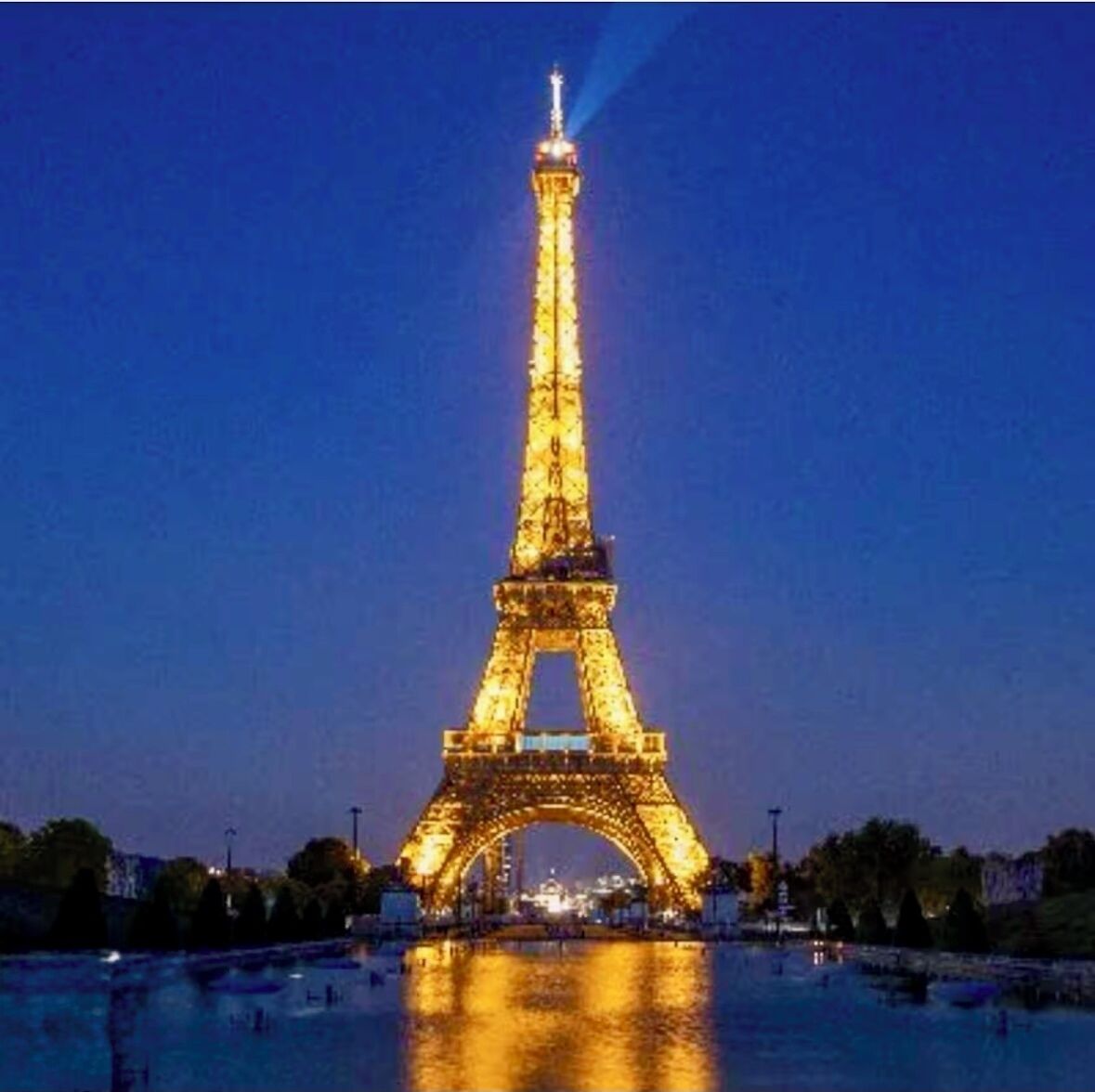 La Torre Eiffel de nit, amb les llums il·luminant-la, reflectint-se a l'aigua.