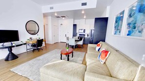 A modern living room with a sofa, a coffee table, and a TV. There is a kitchen area in the background with a dining table and chairs.