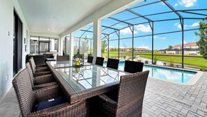 A covered outdoor dining area with a glass table and wicker chairs, overlooking a swimming pool and a golf course. -  9BR Luxury Disney Villa Best Location @ Storey Lake Pool, Theater Sleeps 20 (Kissimmee)