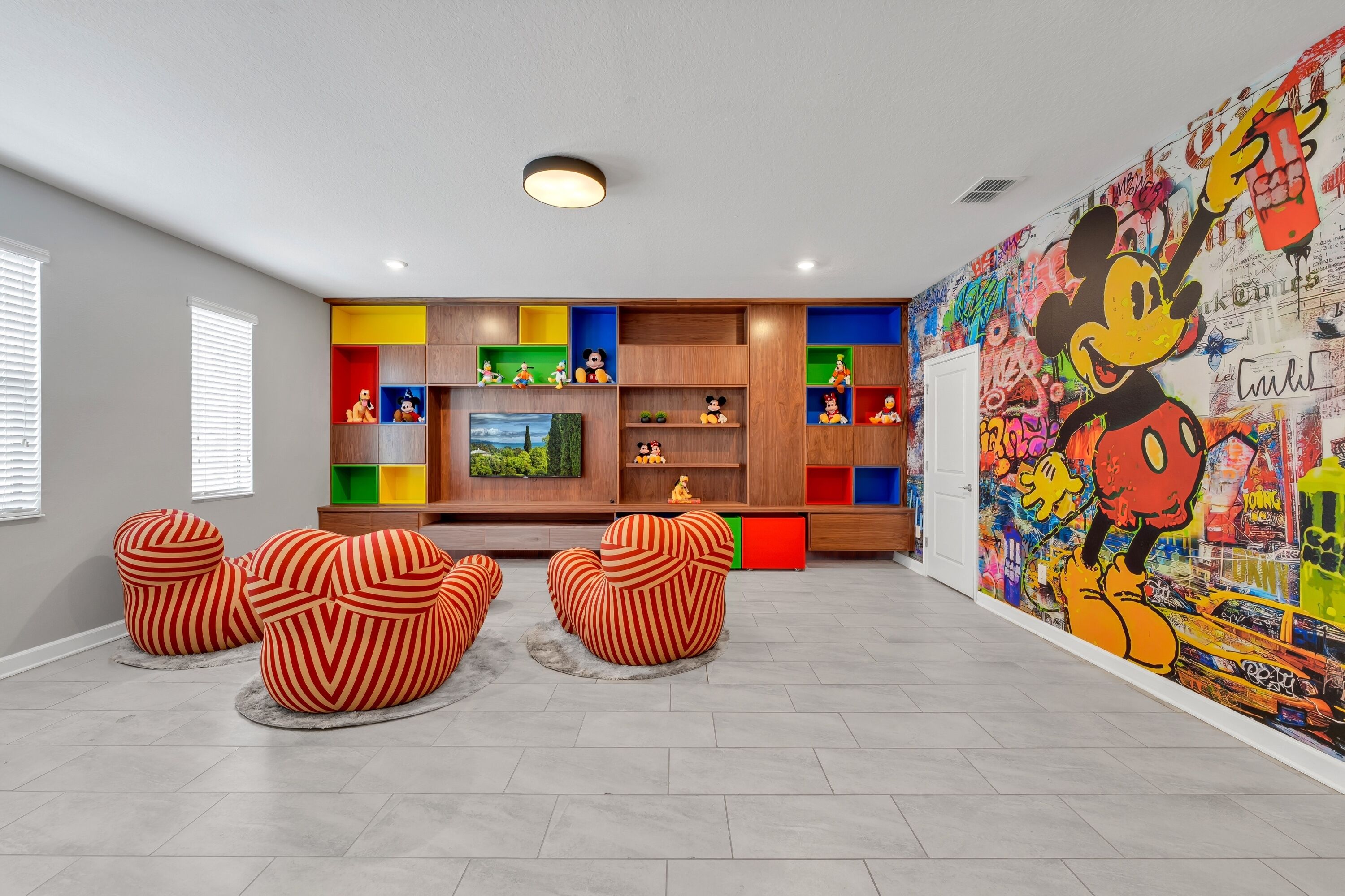 A modern living room with a colorful entertainment unit, a large Mickey Mouse mural, and striped ottomans.