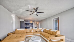 Una sala moderna con un sofá seccional, un ventilador de techo y una cocina al fondo.